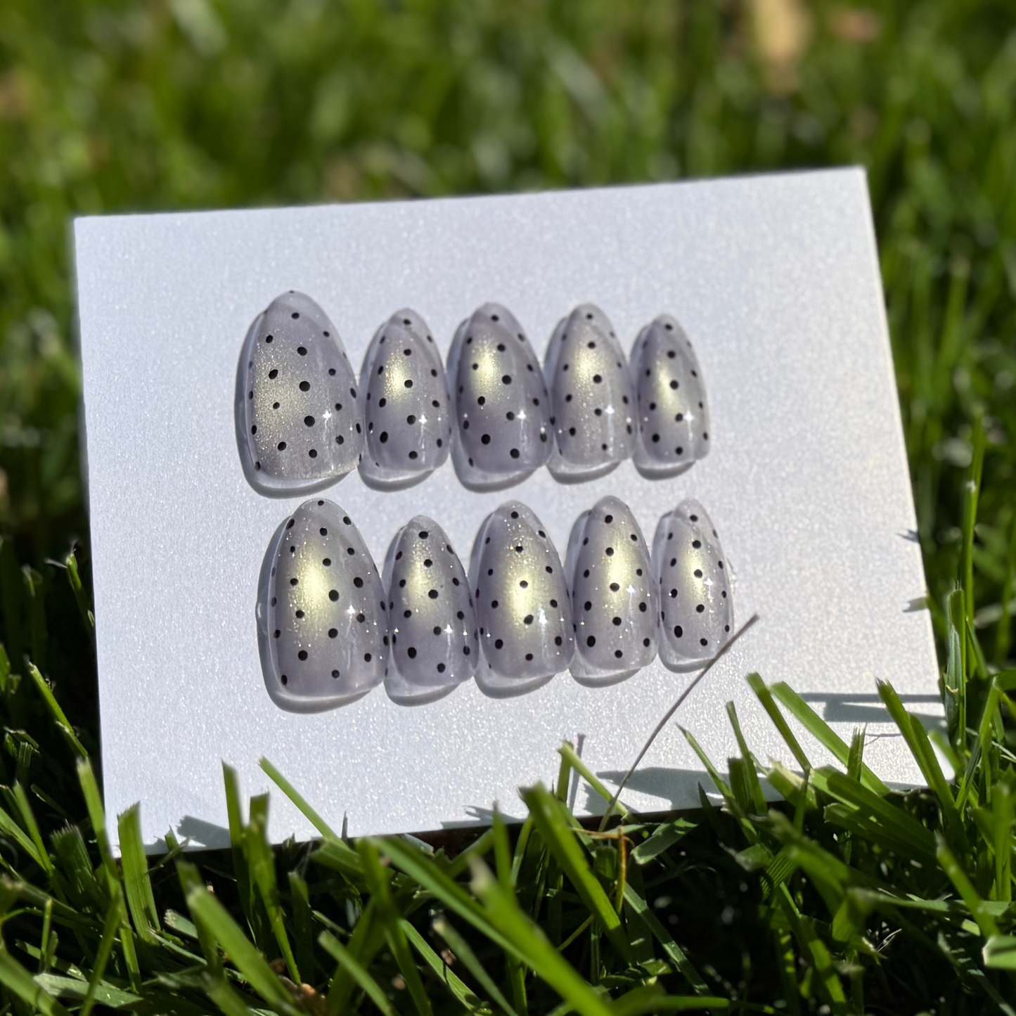 Press-on nails with black polka dots on a white card sitting in the grass
